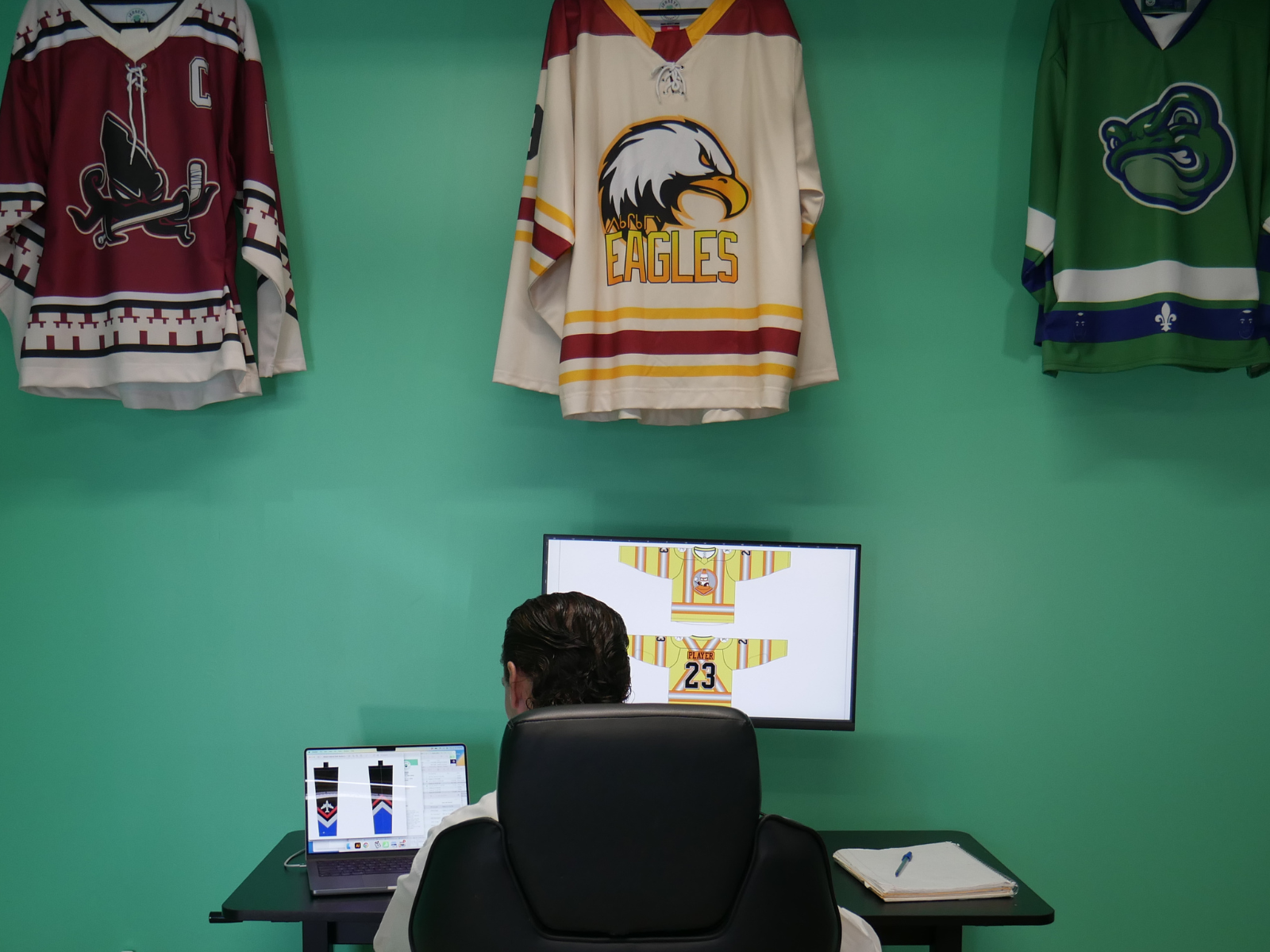 Hockey jersey designer at their desk, on a desktop computer, designing custom jersey artwork with custom jerseys hanging above them on the wall.